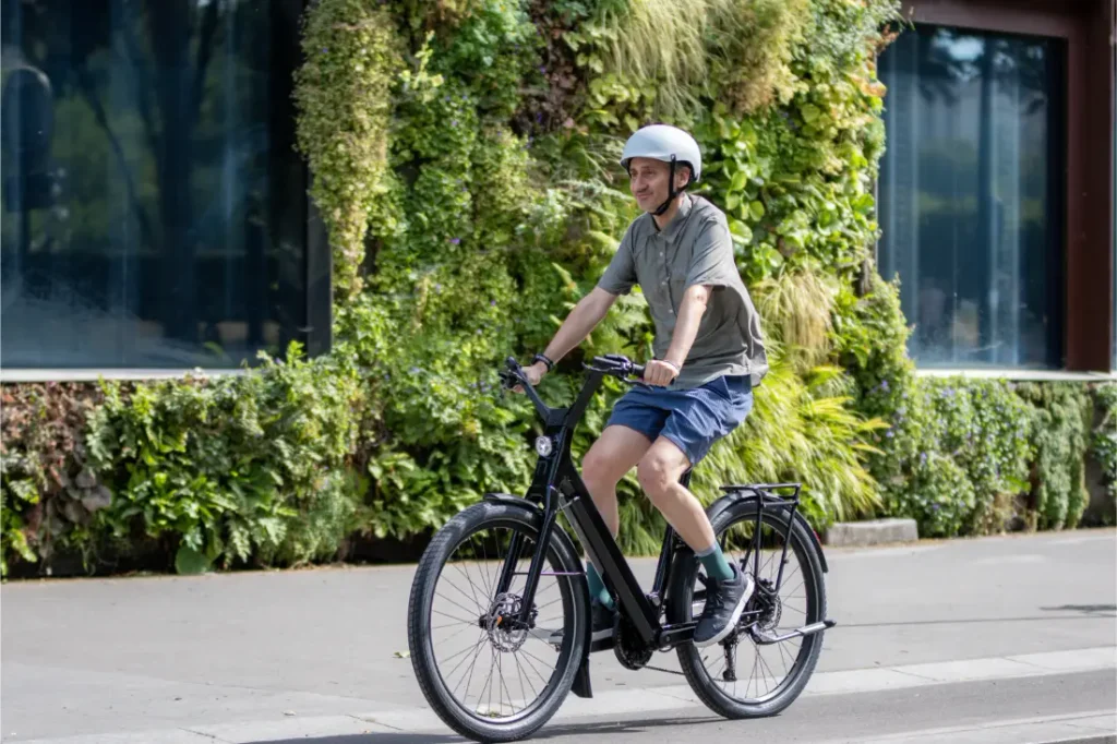 Una persona con casco circula en una bicicleta eléctrica «Moustache Lundi 27» por una calle de la ciudad, con un jardín vertical verde como telón de fondo.