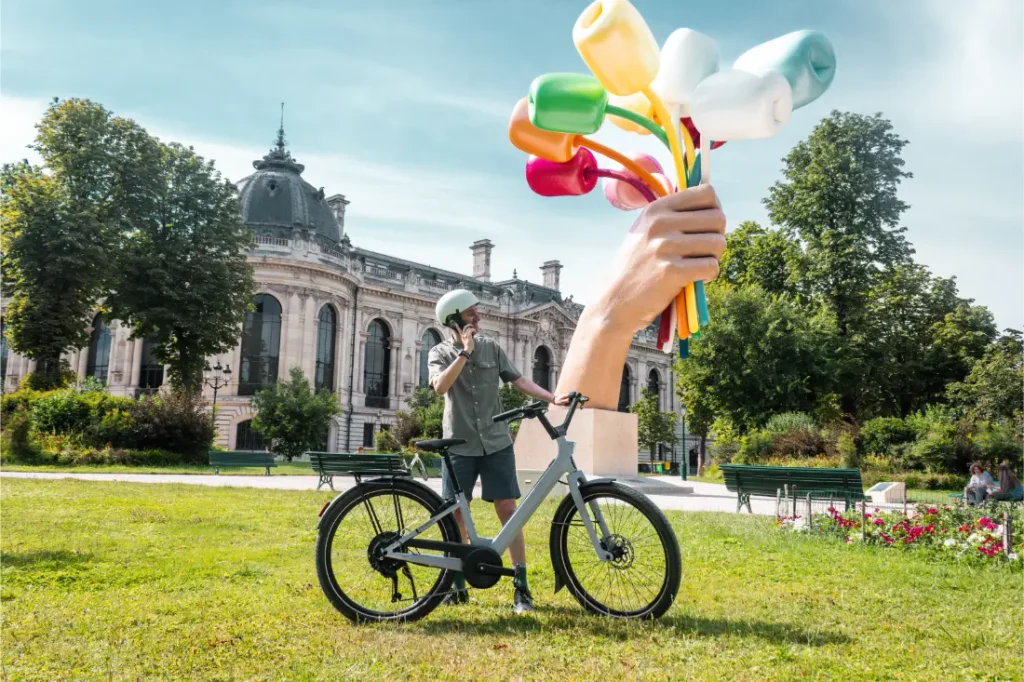 Una persona está de pie junto a una bicicleta «Moustache Lundi 27» sobre la hierba, hablando por teléfono, cerca de una escultura de una mano gigante que sostiene objetos de colores, con un edificio histórico al fondo, lo que transmite el animado ambiente de la ciudad.