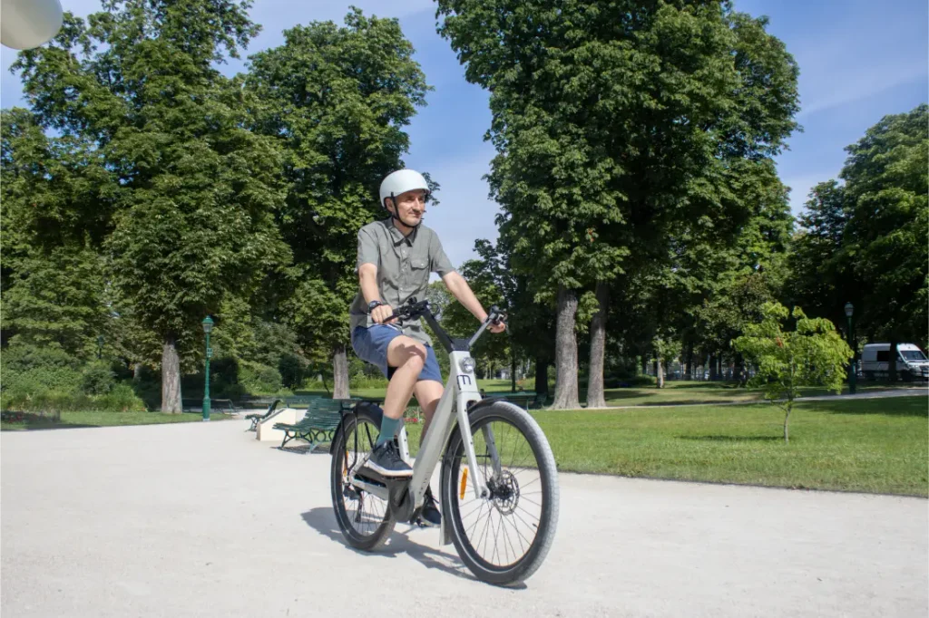 Una persona con casco va en una bicicleta eléctrica «Moustache Lundi 27» por un sendero de un parque urbano, rodeada de árboles y césped verde en un día soleado.