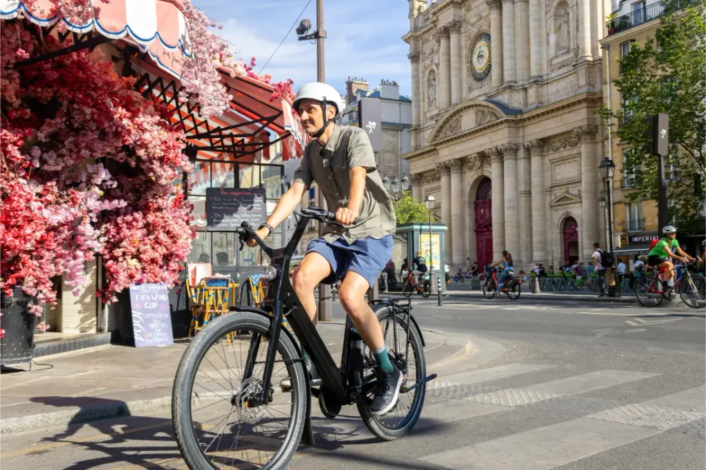 Una persona que lleva casco va en una bicicleta «Moustache Lundi 27» por una calle de la ciudad, cerca de un edificio con grandes columnas y una cafetería adornada con flores rosas.