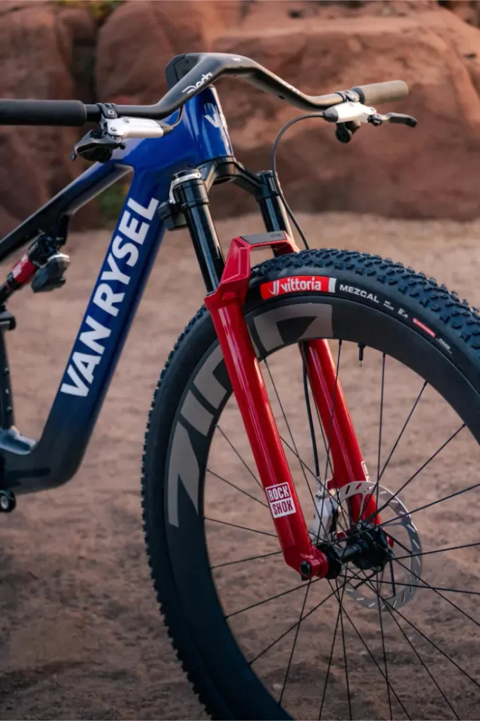 Bicicleta de montaña Van Rysel con cuadro azul, horquilla delantera RockShox roja y neumáticos con tacos.