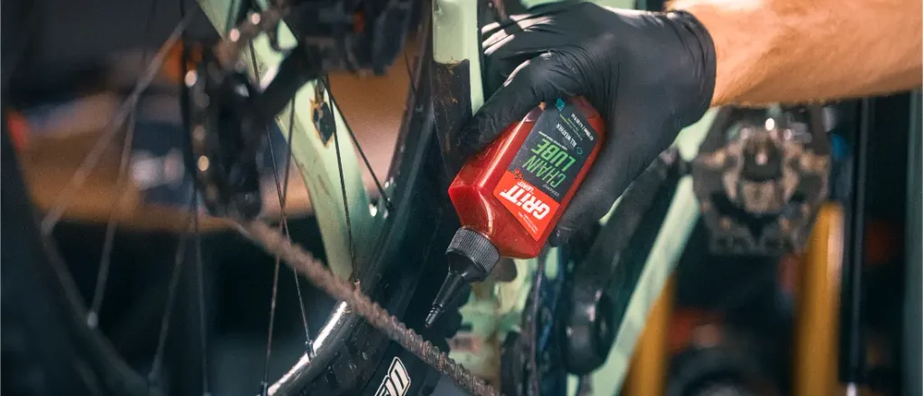 Una mano con un guante negro aplicando lubricante para cadenas Grit, apto para cualquier clima, a la cadena de una bicicleta.