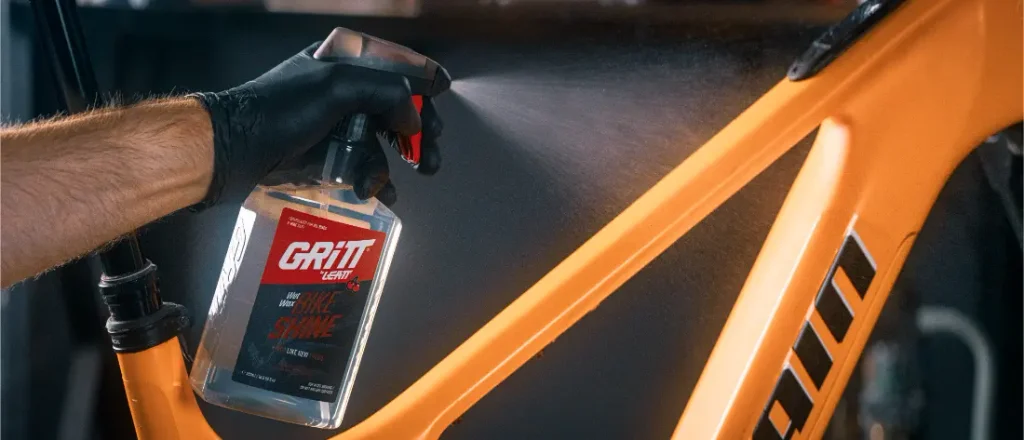 Una mano con un guante negro pulveriza el producto de limpieza «Gritt Bike Shine» sobre el cuadro naranja de una bicicleta.