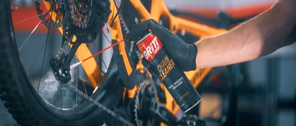 Una mano con un guante negro aplica un bote de spray del limpiador de cadenas Gritt sobre la transmisión de una bicicleta.