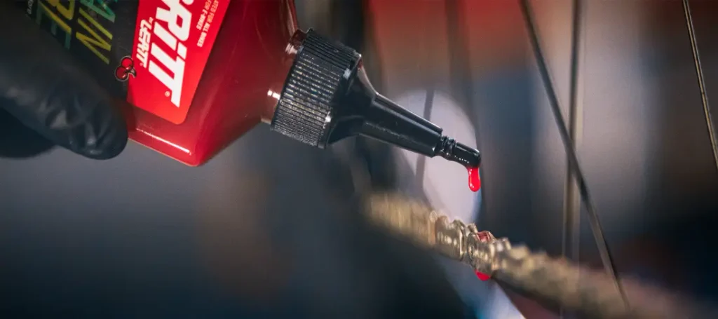 Una mano enguantada que aplica lubricante rojo para cadenas de bicicleta a una cadena desde un frasco dosificador.