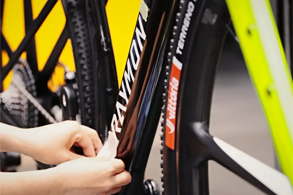 Unas manos colocan una lámina adhesiva protectora en el cuadro de una bicicleta de gravel RAYMON.