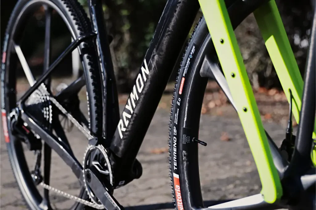 Primer plano de un cuadro negro de bicicleta de grava RAYMON con horquilla delantera de color verde brillante y neumáticos Vittoria con banda de rodadura rugosa.