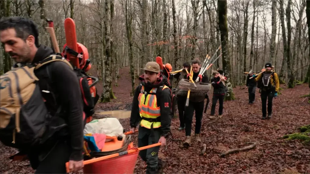 Un grupo de personas que hacen senderismo por un bosque, cargando con herramientas y mochilas.
