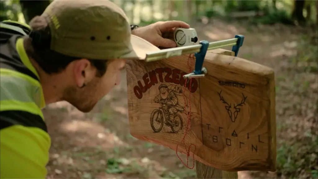 Una persona utiliza una herramienta de nivelación para alinear una señal de senderismo de madera en la que aparecen la ilustración de un ciclista y los textos «Olentzero» y «Trail Born».