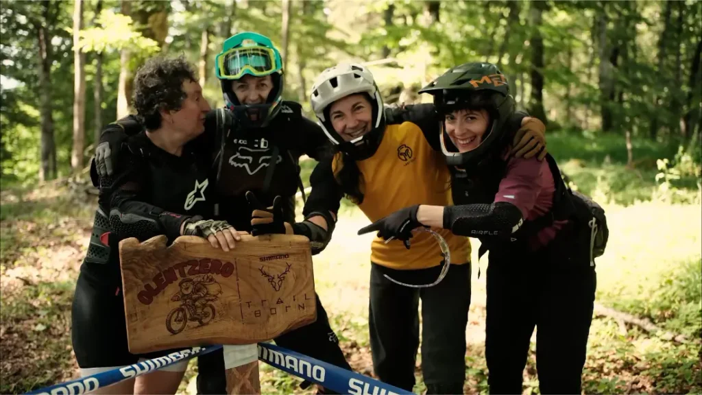 Cuatro ciclistas de montaña de pie juntos en un bosque, sosteniendo una señal de madera en la que se lee «Olentzero Trail Born».