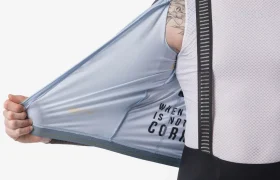 Una persona vestida con ropa de ciclismo sostiene abierto un chaleco Castelli Transition de color azul claro, dejando a la vista una camiseta interior de malla blanca y tirantes negros, perfectos para la temporada de manga larga o para combinar con tu maillot Corretto LS favorito.