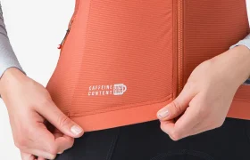 Persona que lleva un chaleco deportivo naranja con cremallera sobre una camiseta Corretto de manga larga con la inscripción «alto contenido en cafeína» cerca del dobladillo, tirando del borde inferior; solo se ven el torso y las manos.