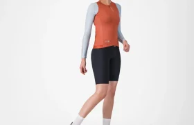 Mujer que lleva un maillot Corretto LS de manga larga en naranja vivo, culotes de ciclismo negros, calcetines blancos y zapatillas deportivas blancas, de pie frente a un fondo blanco liso.