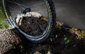 Primer plano de una rueda de bicicleta de montaña llena de barro, con el logotipo amarillo de Pirelli y la inscripción «Scorpion neumáticos MTB», apoyada sobre tierra, piedras y trozos de corteza en el interior de una habitación.