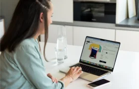 Una mujer está sentada a una mesa utilizando un ordenador portátil para comprar por Internet un maillot de ciclismo amarillo y azul. Sobre la mesa hay un vaso, una jarra de agua y un smartphone.