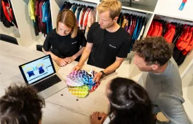 Cuatro personas observan un ordenador portátil en el que se muestran diseños gráficos a todo color y comentan muestras de tejidos en una tienda de ropa o un estudio de diseño, con diversas prendas colgadas en percheros al fondo.