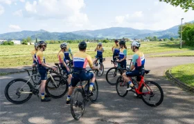 Ocho ciclistas, vestidos con uniformes a juego, se reúnen con sus bicicletas en un camino asfaltado, rodeados de campos y colinas bajo un cielo parcialmente nublado.
