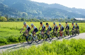 Nueve ciclistas, vestidos con equipación a juego, pedalean en fila por un camino asfaltado que atraviesa un prado verde, con las montañas al fondo.