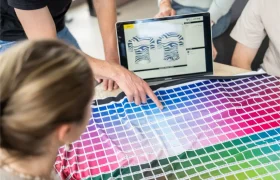Un grupo examina una carta de muestras de telas de colores, mientras una persona señala un color y un ordenador muestra diseños de camisetas en la pantalla.