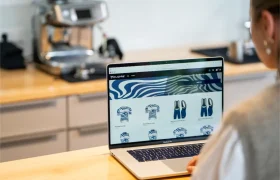 Una persona que navega por una tienda online en la que se muestran prendas deportivas con estampados azules y blancos en un ordenador portátil, en una cocina con una cafetera al fondo.