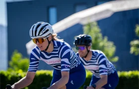 Dos ciclistas, con maillots a rayas azules y blancas a juego y cascos, pedalean al aire libre en un día soleado, con un edificio borroso y vegetación al fondo.