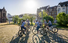 Un grupo de ciclistas con maillots a juego se reúne en círculo en una plaza empedrada, con la luz del sol y los edificios históricos como telón de fondo.