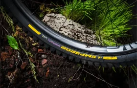 Un primer plano de una rueda de bicicleta de montaña con la inscripción «EMP ERROR» sobre un terreno embarrado, con hierba verde y una roca cerca, en el que se muestran los neumáticos Scorpion MTB de Pirelli, que ofrecen una tracción superior.