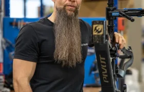 En un concurrido taller de Central Europea de Pivot, una persona con barba larga y gorra hacia atrás permanece de pie, con confianza, sosteniendo un cuadro de bicicleta.