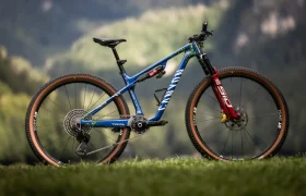 Una bicicleta de montaña con suspensión total, cuadro azul, horquilla roja, neumáticos de color tostado y la marca "Canyon", colocada al aire libre sobre hierba con un fondo verde difuminado, igual que la que monta Jenny Rissveds.