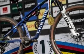 Primer plano de una bicicleta de montaña con una medalla de oro colgada del manillar, delante de un fondo de podio y un cartel con el primer puesto, celebrando la victoria de Jenny Rissveds.