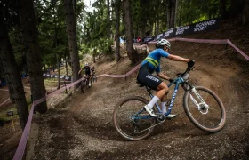 Una ciclista con un maillot de Suecia, posiblemente inspirado en Jenny Rissveds, sube por un sendero de tierra a través de un bosque, seguida de otros ciclistas, durante una carrera de bicicleta de montaña.