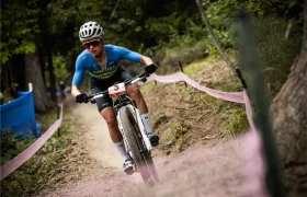 Una ciclista con maillot sueco, parecida a Jenny Rissveds, monta en bicicleta de montaña por un sendero de tierra a través de una zona boscosa, con casco y gafas de sol.