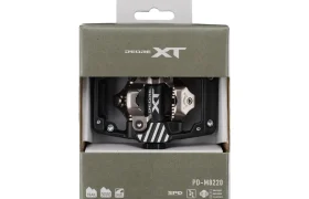 Un pedal de bicicleta de montaña Shimano DEORE XT PD-M8120 SPD Gravel en su embalaje original, visible a través de una ventana transparente en la parte delantera de la caja; unos pedales ideales para el trail y las aventuras todoterreno.