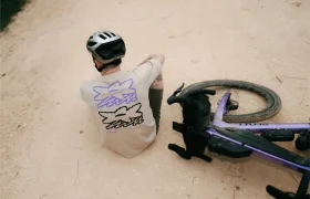 Una persona que lleva casco y una camiseta estampada está sentada en un camino de grava junto a una bicicleta SCOTT Addict Gravel 2027 de color morado, que yace tumbada de lado.