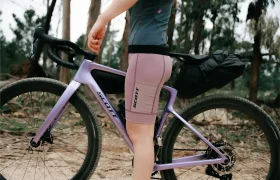 Una persona con ropa de ciclismo posa al aire libre junto a una bicicleta SCOTT Addict Gravel 2027, versátil y cómoda, equipada con una mochila trasera. Al fondo se ven árboles y un suelo de tierra.
