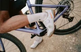 La pierna y el pie de un ciclista, calzado con unas zapatillas blancas de ciclismo Scott, descansan sobre el pedal de una bicicleta morada SCOTT Addict Gravel 2027, conocida por ser polivalente y cómoda, mientras que el otro pie está apoyado en el suelo.
