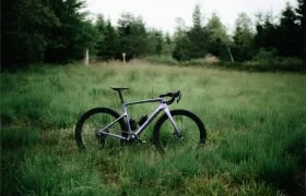 Una SCOTT Addict Gravel 2027 de color morado, una bicicleta polivalente con manillar curvo y neumáticos gruesos para grava, se encuentra sobre la hierba verde en un claro rodeado de árboles.