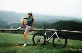 Una persona con ropa de ciclista y casco se apoya en una valla de madera junto a una bicicleta «cómoda» y polivalente SCOTT Addict Gravel 2027, con vistas a un paisaje cubierto de hierba y colinas al fondo.