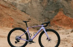 Una bicicleta gravel polivalente, la SCOTT Addict Gravel 2027, en un impresionante color morado, con neumáticos y manillar negros, se alza sobre un terreno arenoso con colinas rocosas al fondo.