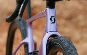 Primer plano de una bicicleta SCOTT Addict Gravel 2027 de color morado, con manillar de carretera y neumáticos de tacos, sobre un fondo difuminado en tonos tierra. Esta bicicleta de gravel polivalente ofrece una experiencia de conducción cómoda en terrenos variados.