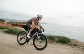 Una persona que lleva casco y gafas de sol monta en una bicicleta gravel polivalente, como la SCOTT Addict Gravel 2027, por un camino de tierra a lo largo de un paisaje costero con acantilados y el mar al fondo.