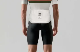 Una persona está de pie mirando hacia otro lado, vistiendo una camiseta de ciclismo Alpinum TotalEnergies blanca y gris con logotipos, pantalones cortos negros, calcetines blancos y zapatillas de ciclismo sobre un fondo liso.