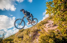 Una persona en bicicleta de montaña se lanza desde un sendero de tierra en un paisaje montañoso, bajo un cielo azul salpicado de nubes blancas.