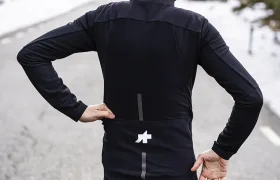 Ciclista con traje negro y casco naranja -con la chaqueta extrema ASSOS- de pie en una carretera de montaña con nieve sobre los hombros, de espaldas a la cámara.