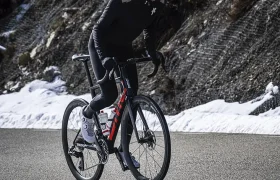 Un ciclista con una chaqueta extrema conduce una bicicleta de carretera cuesta arriba por una carretera asfaltada con nieve a ambos lados y una ladera rocosa y nevada al fondo.