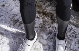 Persona vestida con leggings negros, zapatillas de ciclismo blancas y una Chaqueta ASSOS EQUIPE RS Johdah Ultraz de pie sobre suelo cubierto de nieve junto a un vehículo rojo.
