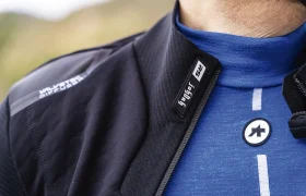 Primer plano de una persona que lleva un maillot de ciclismo azul con el logotipo visible y una chaqueta ASSOS Johdah Ultraz S11 con cremallera parcial, mostrando el texto "SPYKER 2S" en el cuello.
