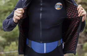 Un hombre con un casco de ciclismo naranja y ropa de ciclismo negra está de pie al aire libre, abriendo la cremallera de su EQUIPE RS Johdah Ultraz chaqueta ASSOS para revelar un forro interior estampado.