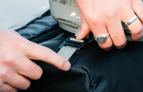 Primer plano de dos manos ajustando una hebilla en un bolso negro ASSOS x Db -perfecto para ciclistas- una persona lleva anillos y un reloj, destacando el elegante sistema de viaje.