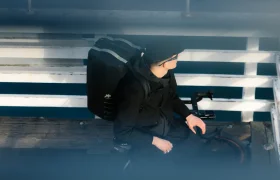 Una persona vestida de negro, con una mochila ASSOS x Db, se sienta en una bicicleta cerca de una barandilla blanca, vista desde arriba a través de unas barras horizontales difuminadas, que captan la esencia de los ciclistas en movimiento y de un innovador sistema de viaje.